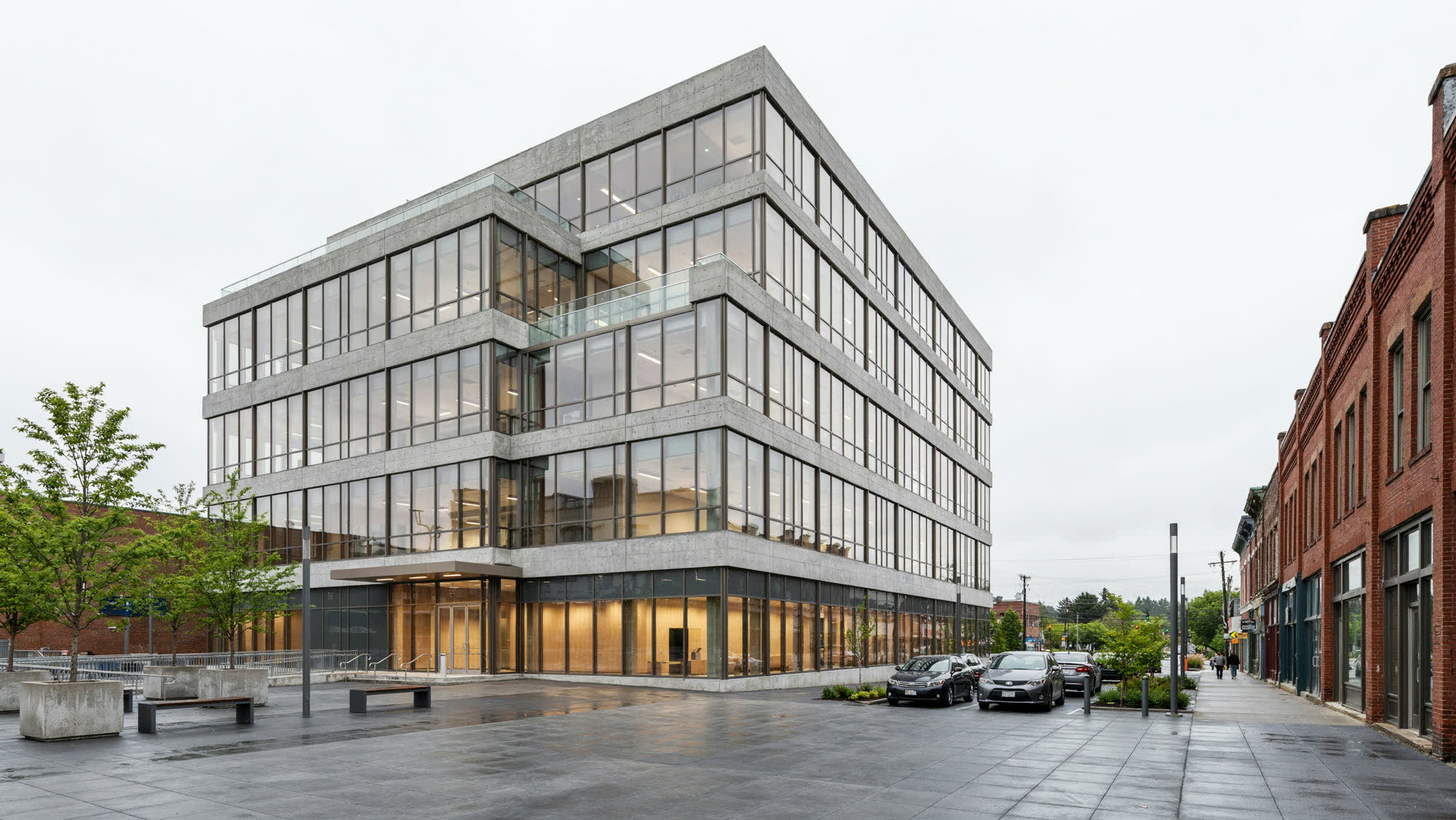 A modern glass and concrete office building on a downtown plaza beside older brick storefronts