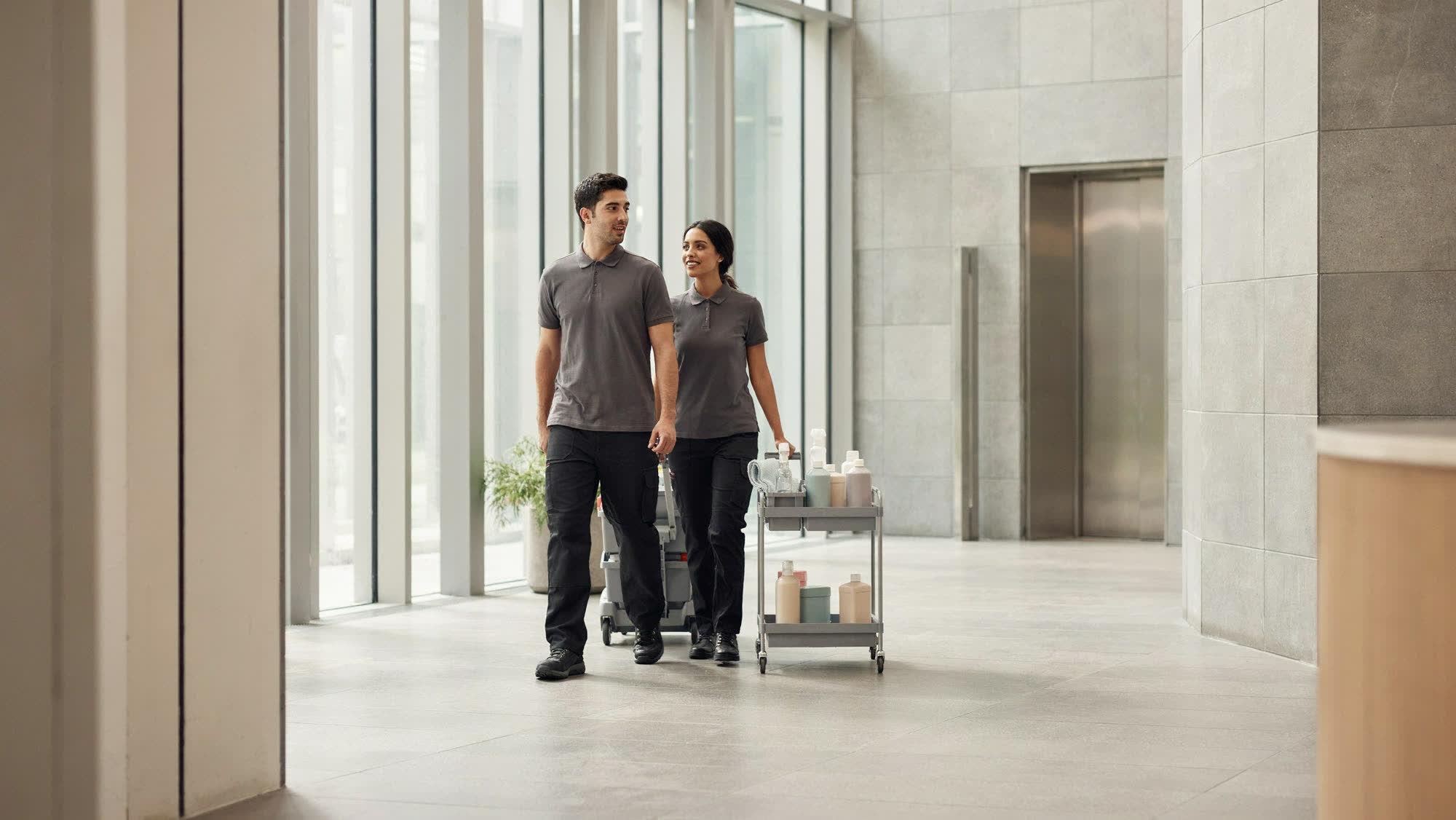Two team members in charcoal polos walking through a bright commercial lobby with a supply cart