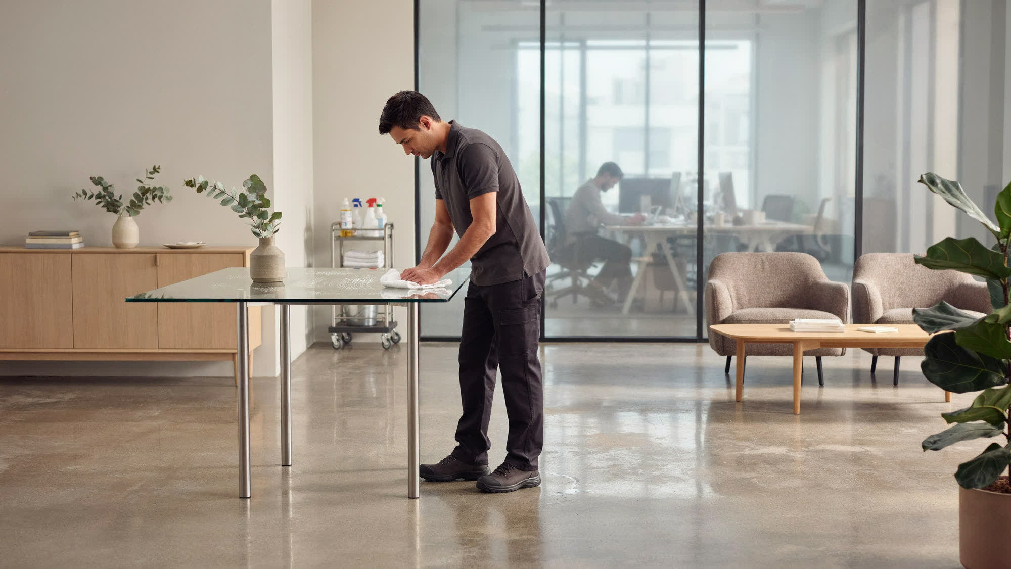 A janitorial team member wiping a glass table in a modern office lounge